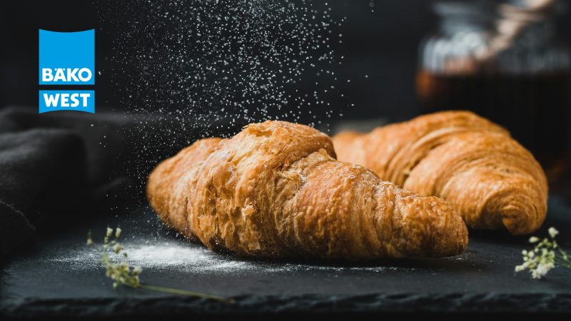 Zwei Goldbraue Croissants werden mit Mehl bestreut, vor dunklem Hintergrund mit dem Logo von BÄKO WEST