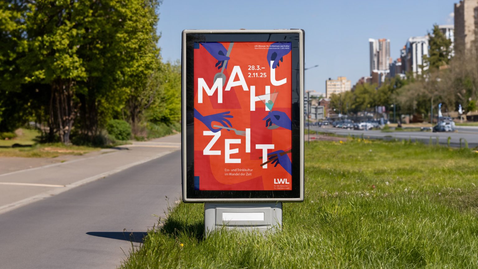 Citylight Poster auf einer Straße mit grünem Rasen. Das Citylight zeigt einen Entwurf für die Archäologieausstellung Mahlzeit im LWL-Archäologiemuseum Herne. Hände schnappen sich die Buchstaben weg.