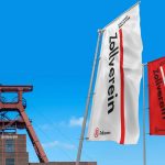 Fahnen mit dem Zollverein-Logo vor dem Fördergerüst des UNESCO-Welterbes Zeche Zollverein unter blauem Himmel.