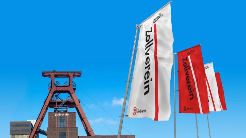 Fahnen mit dem Zollverein-Logo vor dem Fördergerüst des UNESCO-Welterbes Zeche Zollverein unter blauem Himmel.